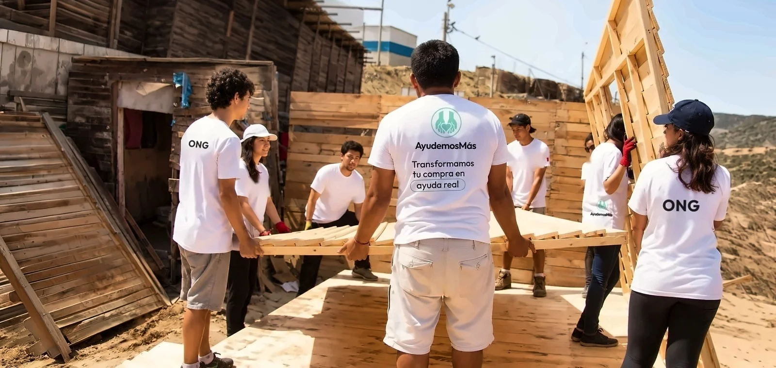 Voluntarios de AyudemosMás con polos blancos levantan juntos un panel de madera para construir una vivienda en una zona arenosa